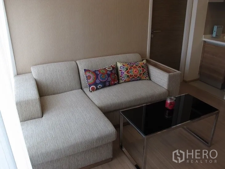 Condominium for rent or sale in Khlong Toei, Bangkok - Living area with light fabric sectional sofa and sleek metal-framed glass coffee table.