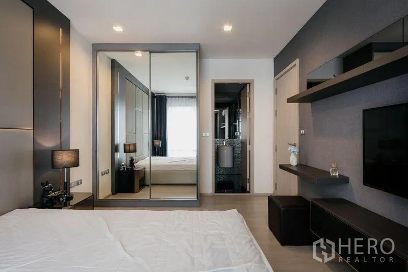 Condominium for rent in Khlong Toei, Bangkok - Bedroom with mirrored wardrobe and doorway to a modern bathroom, with shelves and TV opposite the bed.