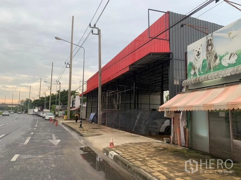 Warehouse for rent in Bueng Kum, Bangkok - Side perspective of high-clearance steel-frame warehouse with red cladding and scaffolding by the curb.