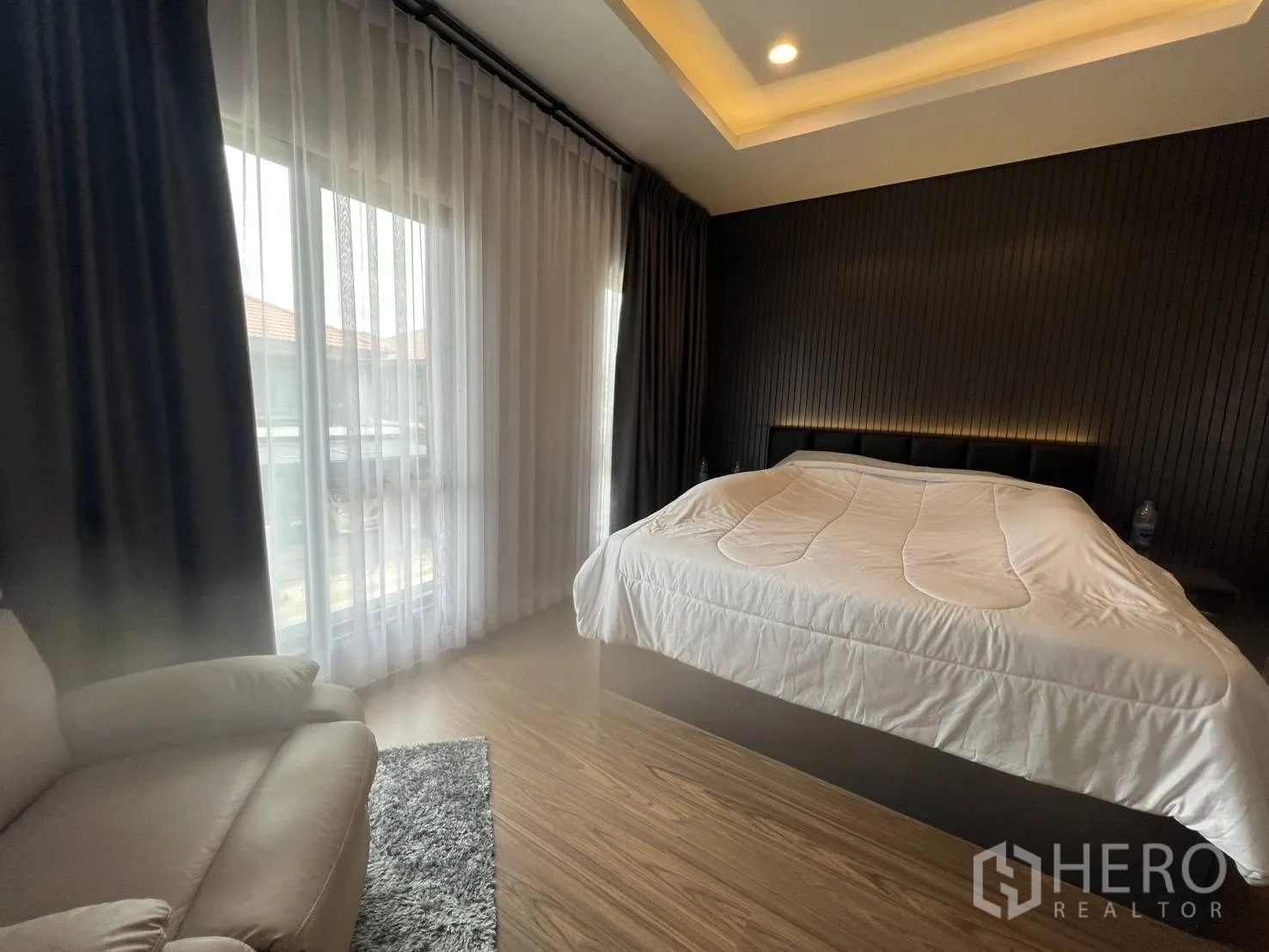Detached House for rent or sale in Lam Luk Ka, Pathum Thani - Master bedroom corner view with floor‑to‑ceiling curtains and wood flooring.