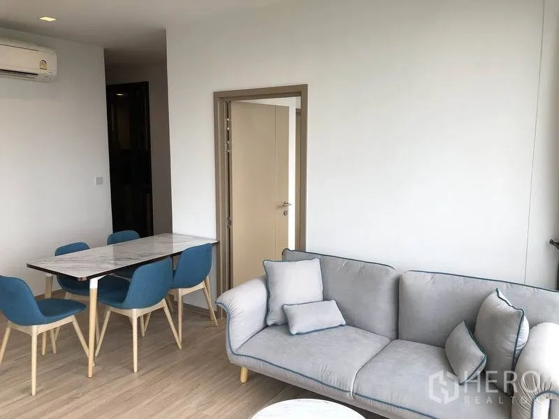 公寓 出租在Watthana, Bangkok - 温馨客厅，灰色沙发与靠垫，旁设紧凑餐桌，极简风格。