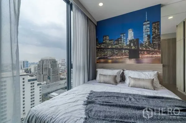 Condominium for rent in Pathum Wan, Bangkok - Corner bedroom with floor-to-ceiling windows, city skyline view and a dramatic cityscape mural behind the bed.