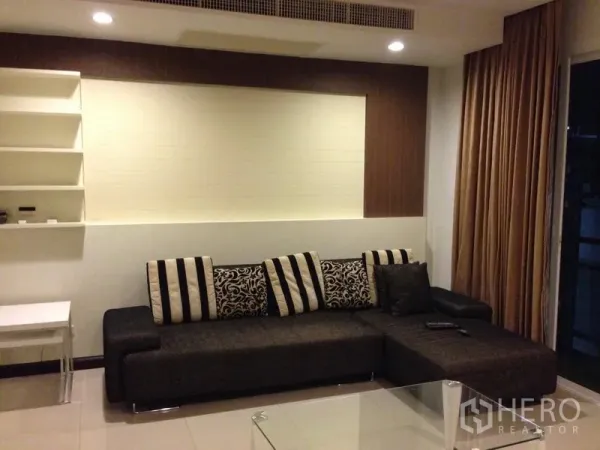 Condominium for rent in Watthana, Bangkok - Cozy lounge with dark L-shaped sofa, striped cushions and built-in shelving next to full-length curtains.
