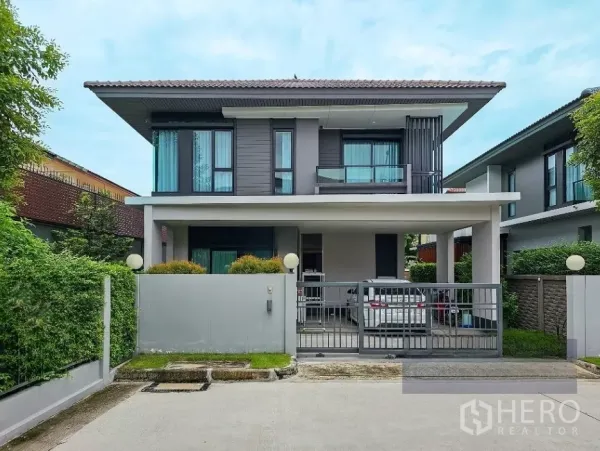 Detached House for sale in Lam Luk Ka, Pathum Thani - Front view of a modern two-storey detached house with gated carport, balcony and hedge fence on a corner plot.