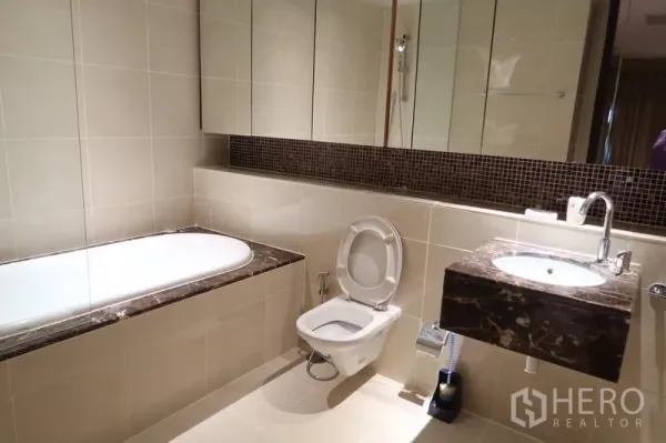 Condominium for rent in Khlong Toei, Bangkok - Modern bathroom with marble-topped vanity, bathtub, large mirror and beige tiles.