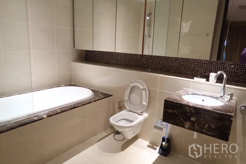 Condominium for rent in Khlong Toei, Bangkok - Modern bathroom with marble-topped vanity, bathtub, large mirror and beige tiles.