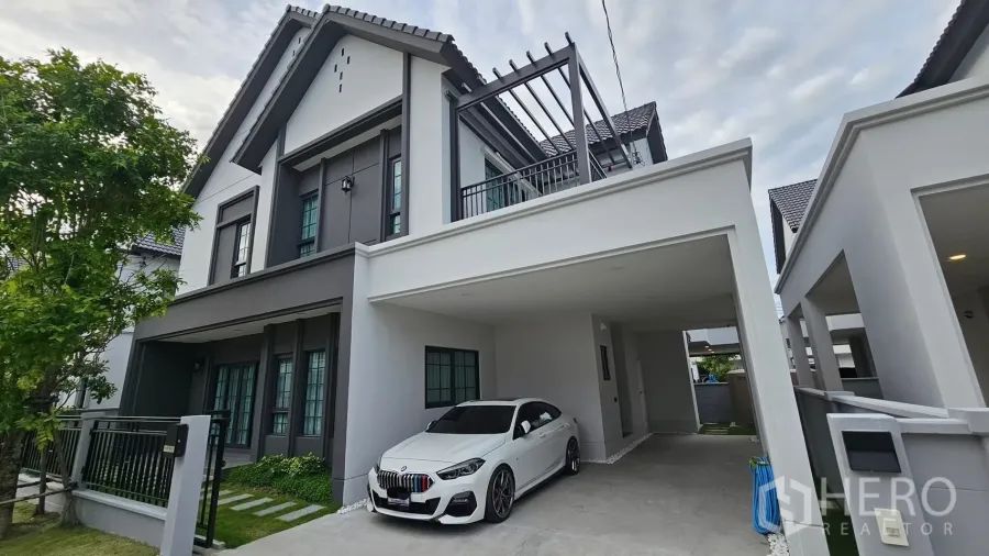 独立式住宅 出租或出售在Bang Phli, Samut Prakan - 现代英式两层独栋外观，带遮棚车位、白色轿车、阳台格栅与景观庭院。