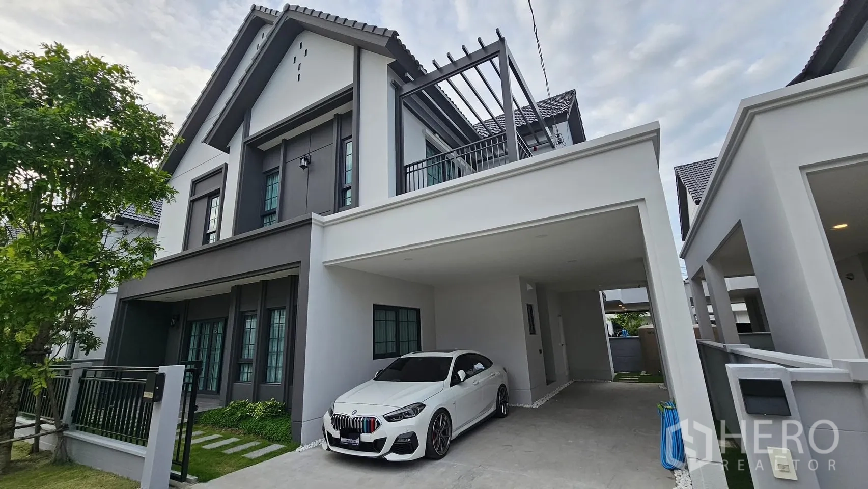 独立式住宅 出租或出售在Bang Phli, Samut Prakan - 现代英式两层独栋外观，带遮棚车位、白色轿车、阳台格栅与景观庭院。