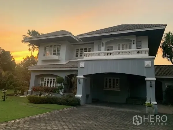 Detached House for rent in Khan Na Yao, Bangkok - Two-storey detached house exterior with covered carport, balcony, and large front yard at dusk.