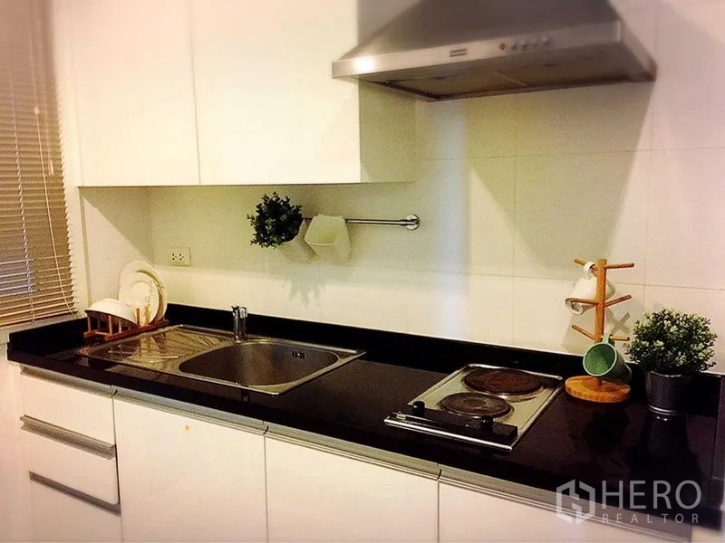 Condominium for rent in Khlong Toei, Bangkok - Modern galley kitchen with black countertop, sink, cooktop, and range hood.