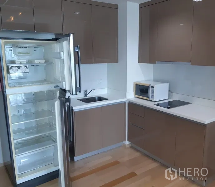 Condominium for rent in Khlong Toei, Bangkok - Modern L-shaped condo kitchen with glossy cabinets, open refrigerator, sink and electric hob in Phra Khanong, Bangkok.