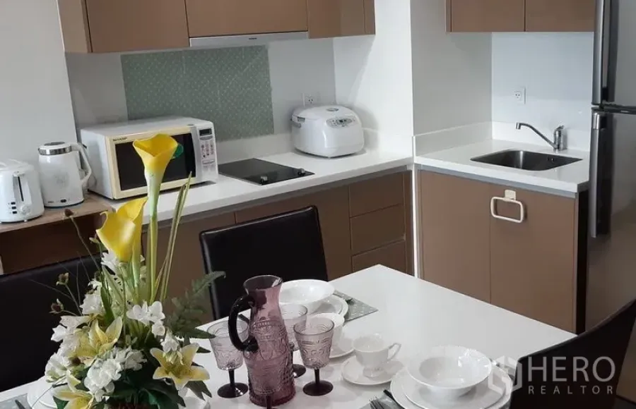 Condominium for rent in Khlong Toei, Bangkok - Open kitchen with white countertops, cooktop, microwave, rice cooker, sink and refrigerator next to the dining table.