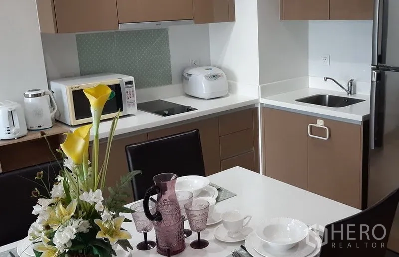 Condominium for rent in Khlong Toei, Bangkok - Open kitchen with white countertops, cooktop, microwave, rice cooker, sink and refrigerator next to the dining table.