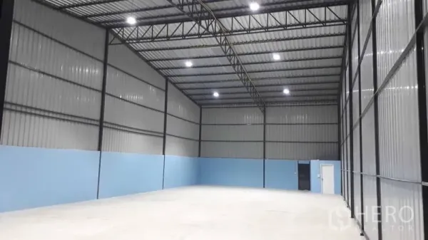 货栈 出租在Pak Kret, Nonthaburi - 开放式仓库区域，高挑层高，蓝色下半墙面，配有人行门和大型滑门。
