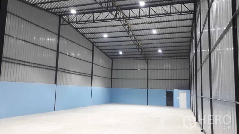 货栈 出租在Pak Kret, Nonthaburi - 开放式仓库区域，高挑层高，蓝色下半墙面，配有人行门和大型滑门。