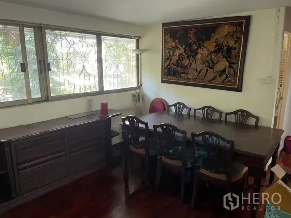 Home Office for rent in Watthana, Bangkok - Dining area with large wooden table, sideboard, window grilles and framed wall art.