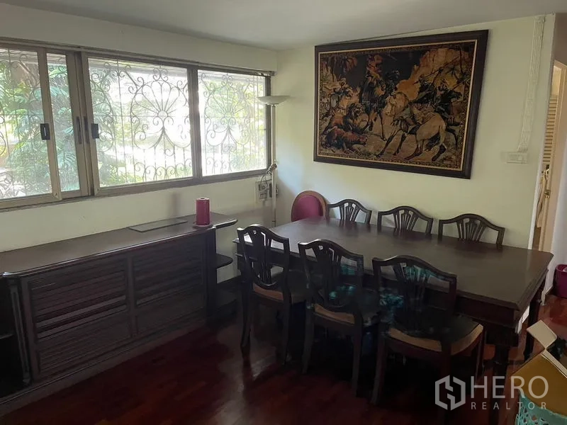 Home Office for rent in Watthana, Bangkok - Dining area with large wooden table, sideboard, window grilles and framed wall art.