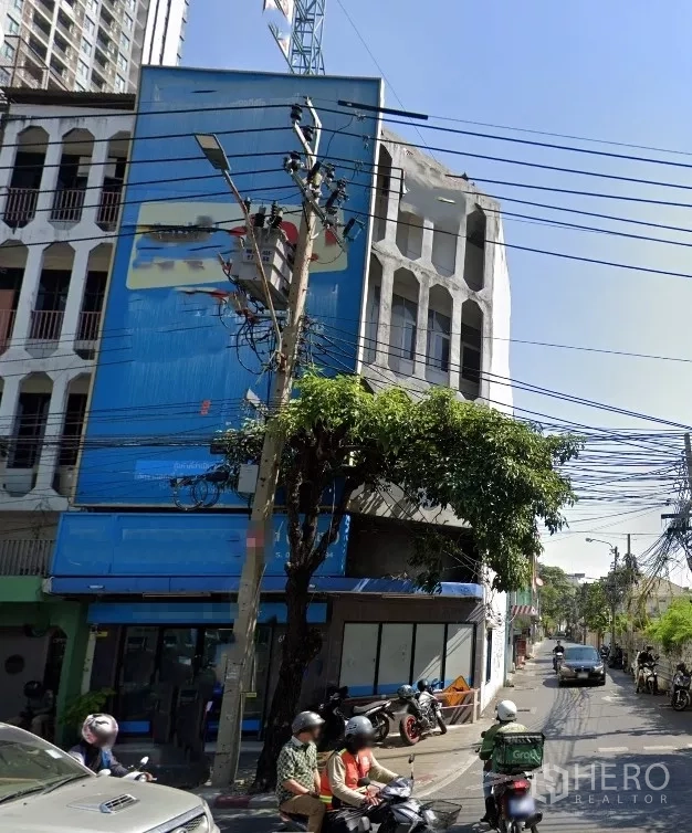 Shophouse for rent in Watthana, Bangkok - Corner 4.5-storey shophouse with glass frontage on a side street, overhead power lines and a shade tree in front near BTS Ekkamai.