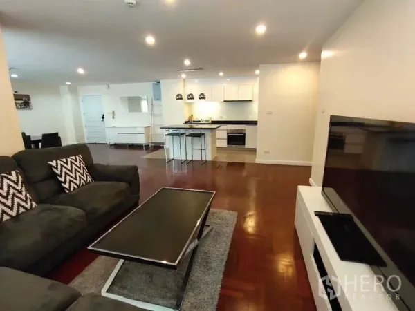 Condominium for rent or sale in Bang Rak, Bangkok - Open‑plan living room with sectional sofa and TV facing a modern white kitchen with island.