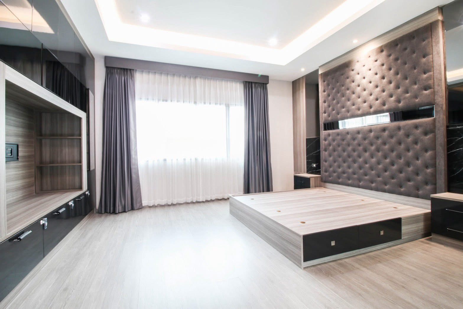 Townhouse for rent or sale in Lat Phrao, Bangkok - Master bedroom with platform bed, tufted feature wall, cove lighting and a large curtained window.