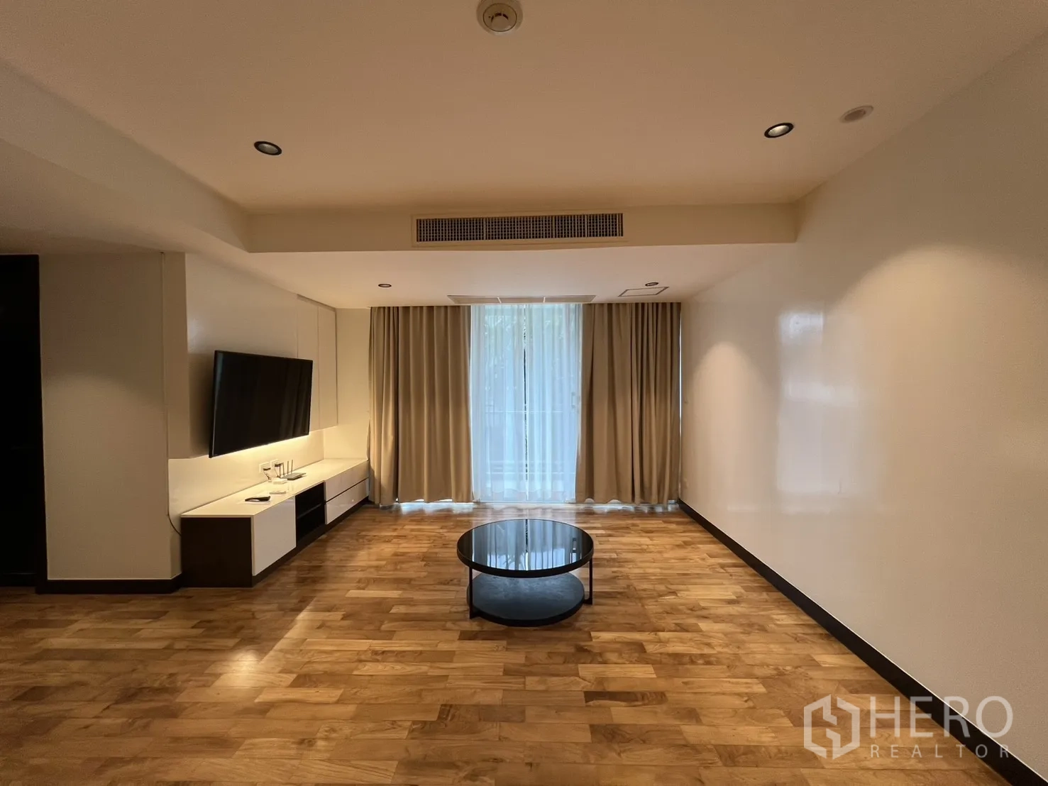 อพาร์ทเมนท์ for rent in Watthana, Bangkok - ห้องนั่งเล่นกว้างพร้อมทีวีติดผนัง ตู้วางสื่อบิวท์อิน โต๊ะกลางทรงกลม และผ้าม่านเต็มความสูง