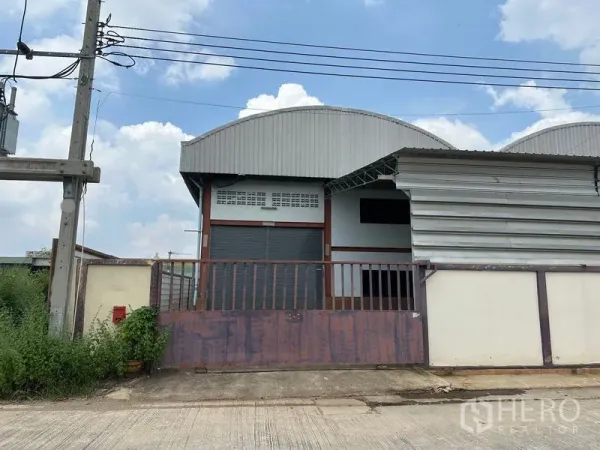 Warehouse for rent in Pak Kret, Nonthaburi - Street-facing warehouse in Pak Kret with arched corrugated metal roof, wide roller shutter and secure gated entrance under a blue sky.