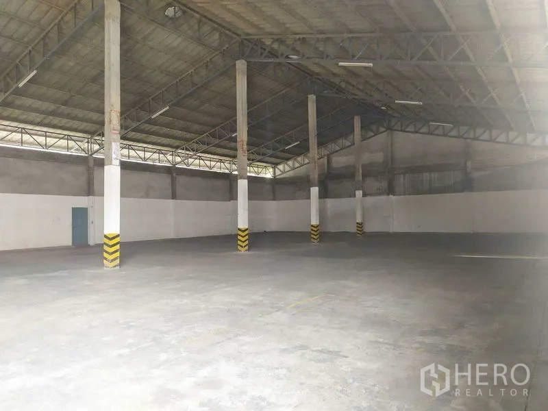 Warehouse for rent in Mueang Samut Prakan, Samut Prakan - Spacious warehouse interior in Mueang Samut Prakan with high steel‑truss roof, safety‑striped columns and durable concrete floor.