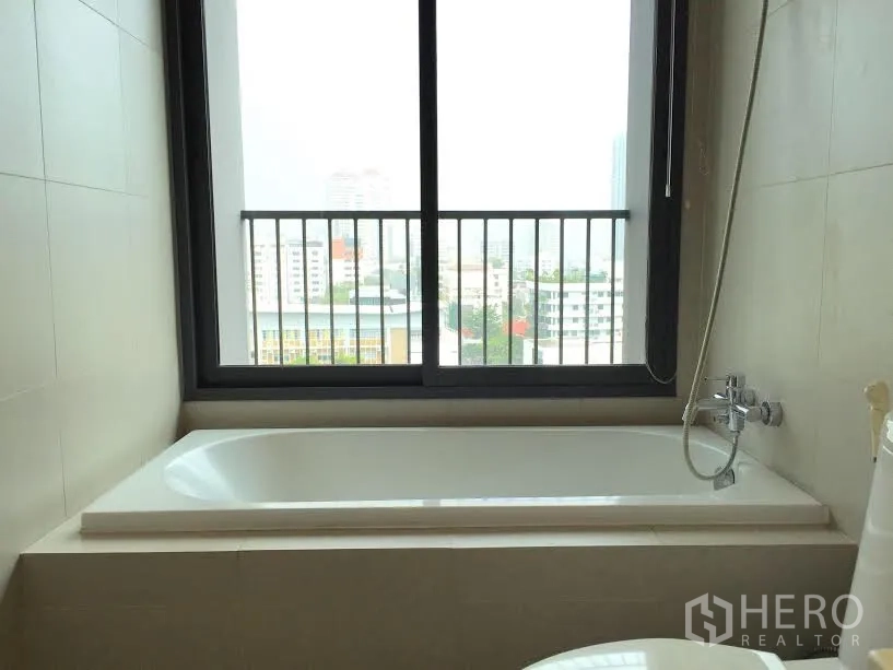 Condominium for rent or sale in Khlong Toei, Bangkok - Bathroom with built-in bathtub beneath a large window offering city views in a Bangkok condominium.