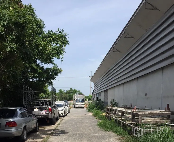Warehouse for rent in Khlong Luang, Pathum Thani - Wide driveway beside a large warehouse with parked pickup trucks and shaded trees, suitable for truck access.
