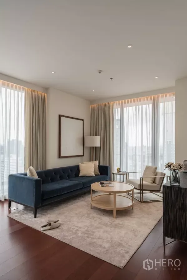 公寓 出租在Watthana, Bangkok - 阳光客厅，蓝色沙发与浅色地毯，通过纱帘可见城市景观。