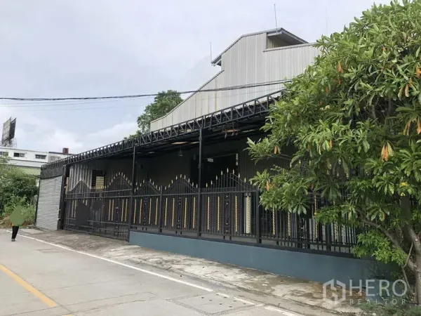 货栈 出租在Bang Phli, Samut Prakan - 金属夹芯板外墙的仓库侧视，装饰围栏与路边树木。