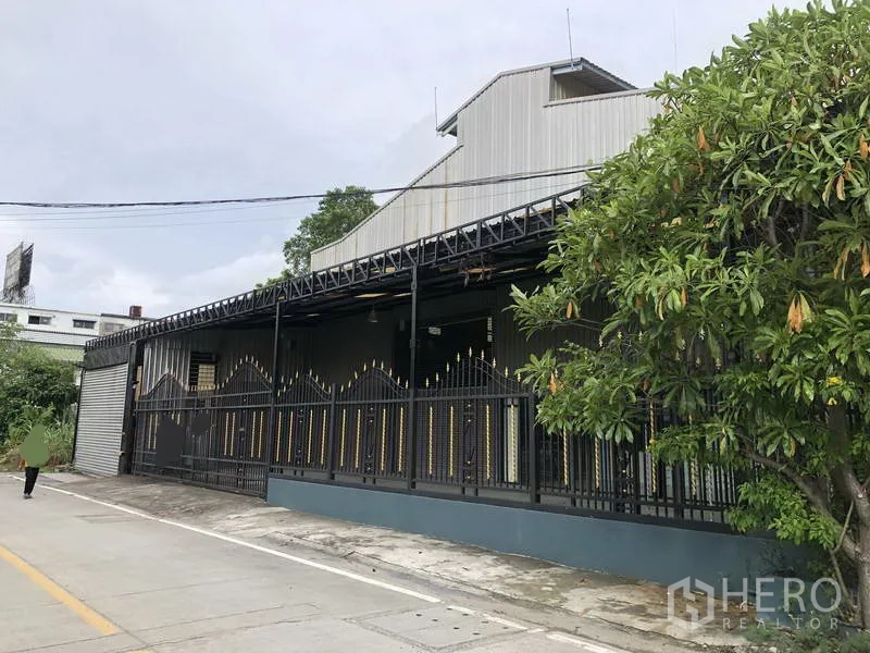 货栈 出租在Bang Phli, Samut Prakan - 金属夹芯板外墙的仓库侧视，装饰围栏与路边树木。