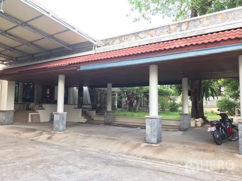独立式住宅 出租在Bang Phli, Samut Prakan - 宽敞有顶车棚，红瓦屋顶与立柱，旁有车道与小草坪。