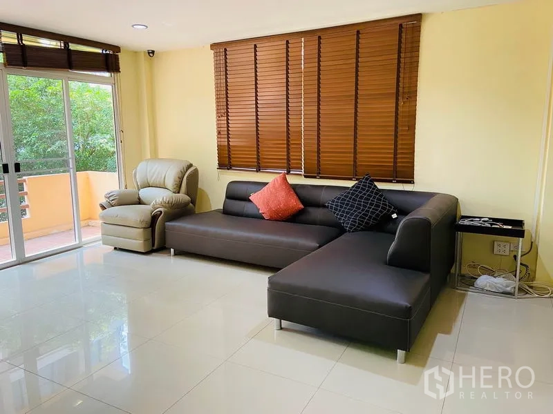 Detached House for rent in Watthana, Bangkok - Living room with L-shaped sofa, armchair, wood blinds and sliding doors to a balcony.