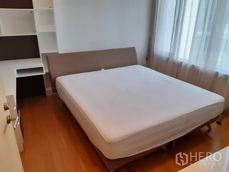 Condominium for rent in Khlong Toei, Bangkok - Cozy bedroom with large bed, built-in shelving, and natural light through sheer curtains.