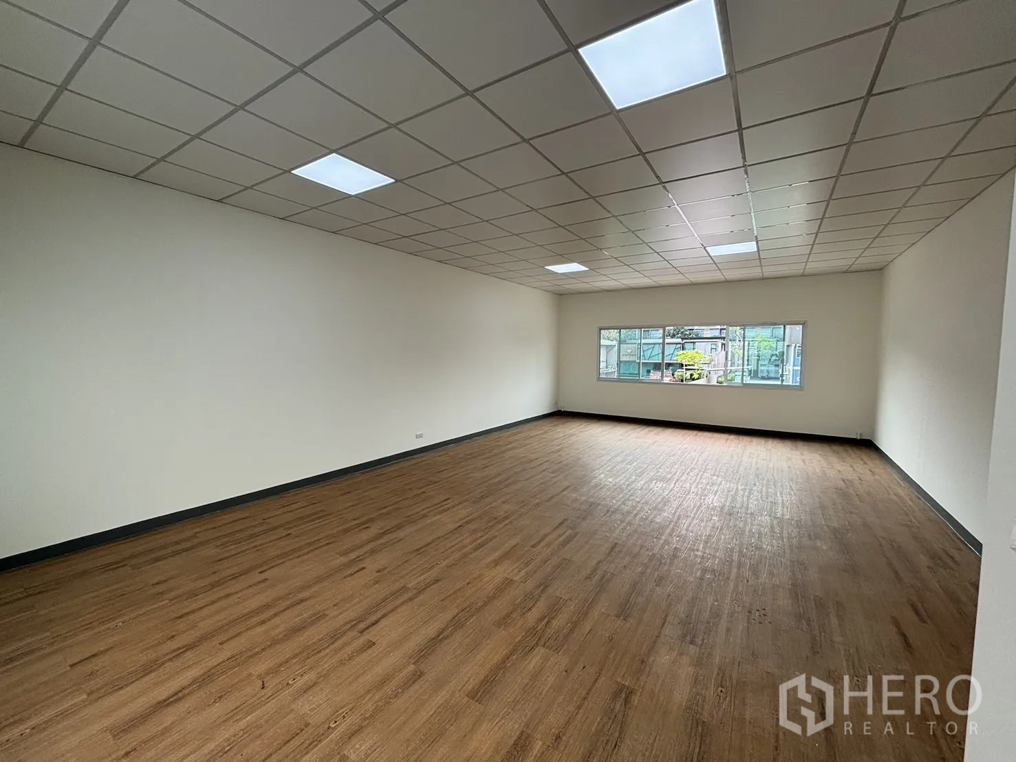 Retail Space for rent - Large empty retail room with wood-look floors, white walls, drop ceiling lights and a window.