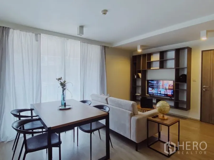 Condominium for rent in Pathum Wan, Bangkok - Bright living and dining area with sofa, four-seat table, built-in shelving and TV, filtered by sheer curtains.