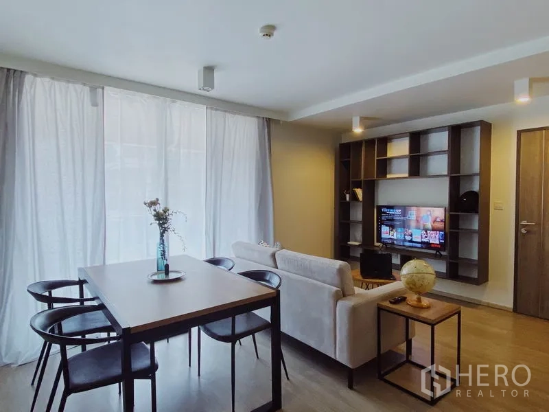 Condominium for rent in Pathum Wan, Bangkok - Bright living and dining area with sofa, four-seat table, built-in shelving and TV, filtered by sheer curtains.