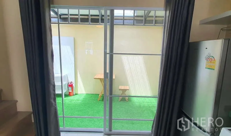 Detached House for rent in Bang Phli, Samut Prakan - View through sliding doors to a covered patio with artificial lawn and small table set.