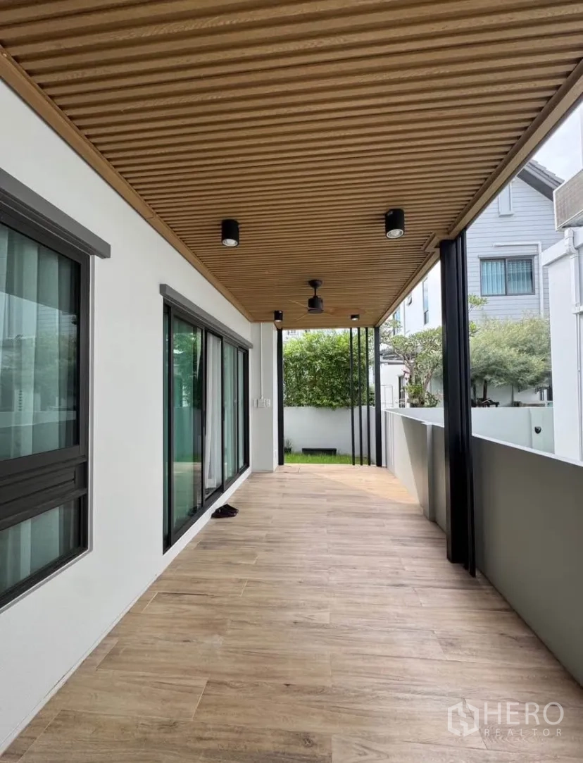Detached House for rent or sale in Lat Krabang, Bangkok - Covered side terrace with wood-look flooring, slatted ceiling, and large sliding glass doors opening to a small lawn.