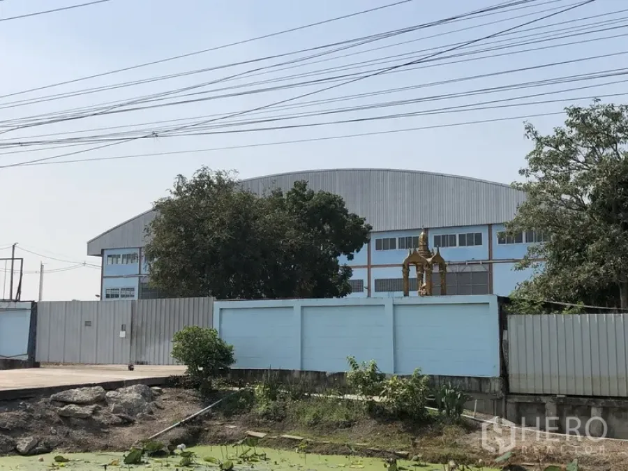 Factory for sale in Lat Lum Kaeo, Pathum Thani - Gated blue factory with arched roof behind a perimeter wall, trees and a canal in an industrial setting.