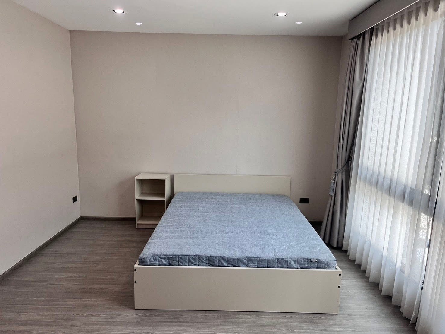 Townhouse for rent or sale in Lat Phrao, Bangkok - Bright bedroom with a simple bed frame beside full-height sheer curtains and grey flooring.