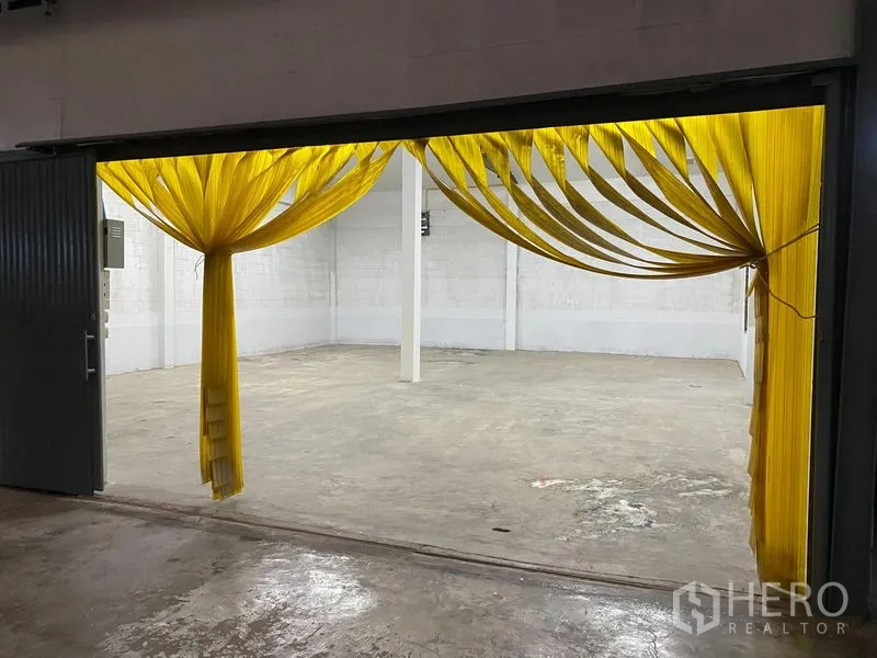 คลังสินค้า ให้เช่า - ประตูโหลดสินค้าขนาดใหญ่พร้อมม่านริ้ว PVC สีเหลือง เผยให้เห็นพื้นที่โกดังพื้นคอนกรีตและเสารับน้ำหนักกลางห้อง
