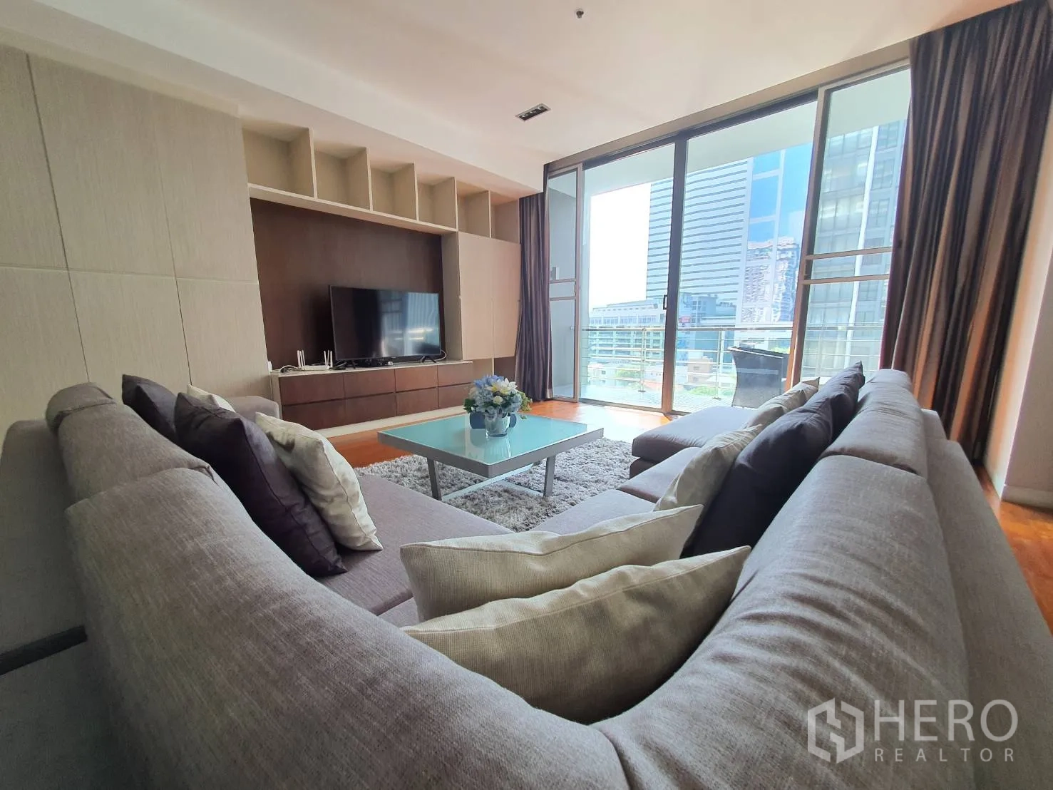 Condominium for rent in Khlong Toei, Bangkok - Bright lounge with grey sectional, glass coffee table and sliding doors to a balcony overlooking high-rises.