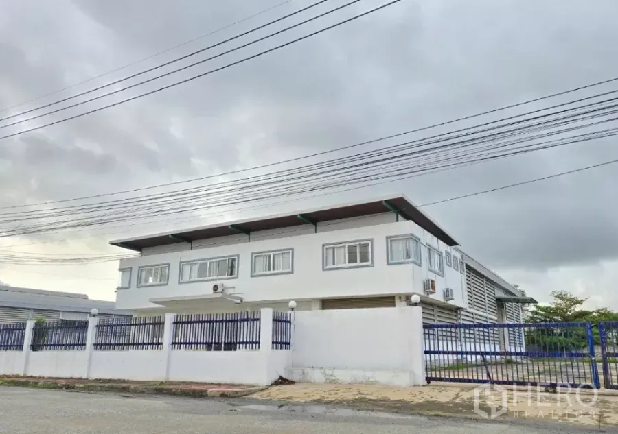 工厂 出售在Mueang Samut Prakan, Samut Prakan - 两层工厂及办公楼外观，蓝色围栏，阴天天空，位于Mueang Samut Prakan。