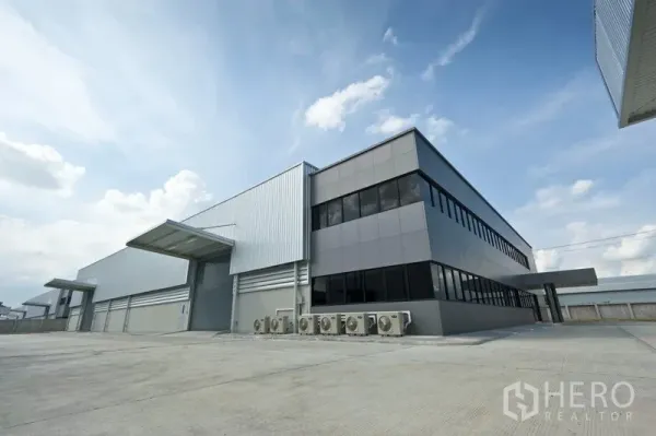 Factory for rent or sale in Samut Prakan - Angle view of two‑storey office and steel factory hall with ample driveway for trucks.