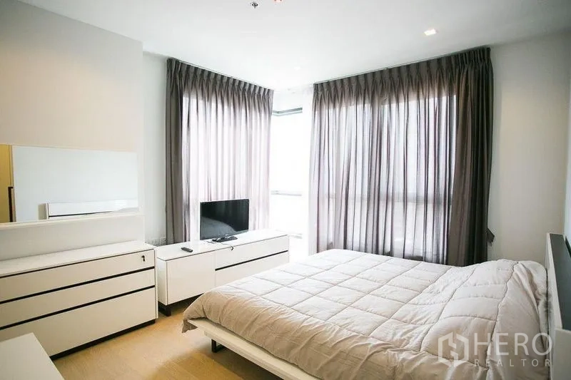 Condominium for rent in Watthana, Bangkok - Primary bedroom with large windows, sheer curtains and sleek white dressers.