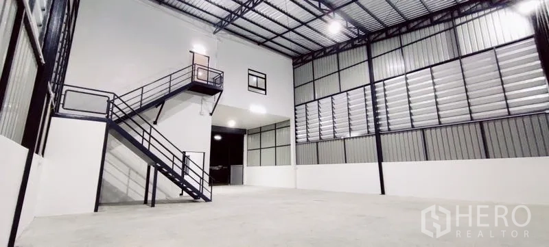 Warehouse for rent in Lam Luk Ka, Pathum Thani - High-bay storage hall with steel staircase to a mezzanine office and bright lighting.