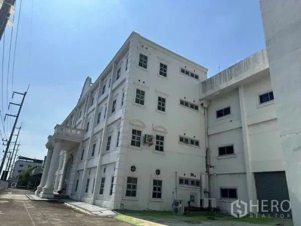 工厂 出租或出售在Thanyaburi, Pathum Thani - 四层白色工厂办公楼外立面，设有古典立柱与侧面装卸区，天空晴朗。