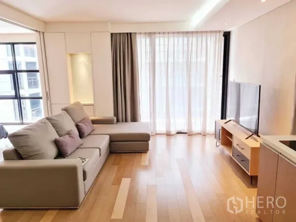 Condominium for rent in Watthana, Bangkok - Open living room with L-shaped sofa, TV console and balcony glass doors with sheer curtains.